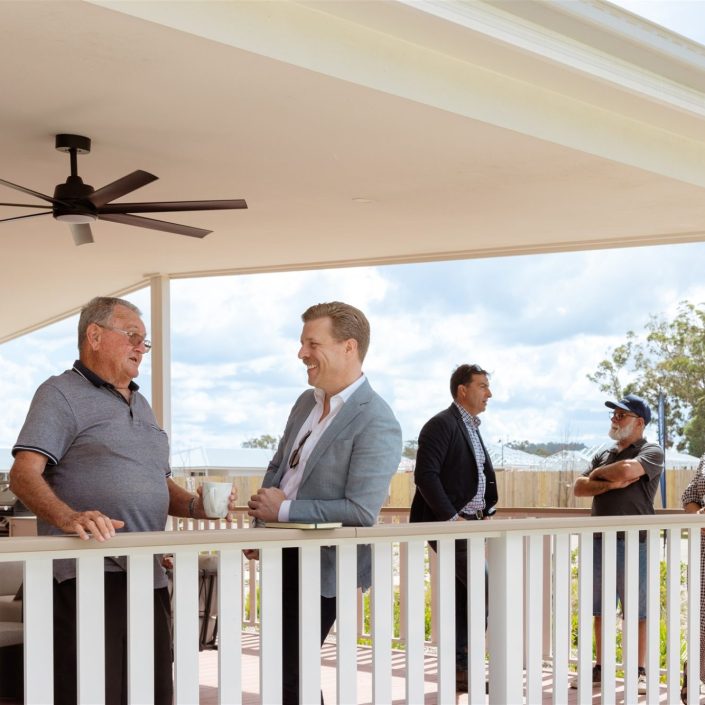 A group from the Northern Rivers over-50s Clubhouse stand and talk on a covered porch; three lean on the railing in conversation whilst others chat in the background, with trees and a cloudy sky behind them.