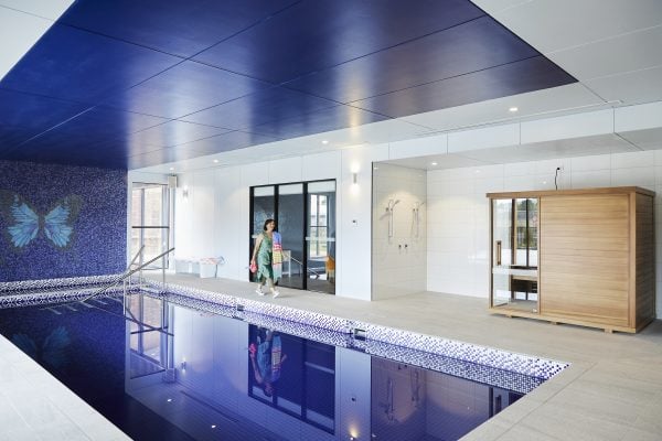 A woman in a swimming costume and towel, enjoying over-50s wellness, walks along the side of a modern indoor pool, featuring a mosaic butterfly design on the wall and a nearby wooden sauna. The bright pool area feels fresh and contemporary.