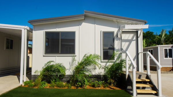 A modern white modular home with large front windows, a small staircase leading to the entrance, green plants in front, and a bright blue sky overhead.