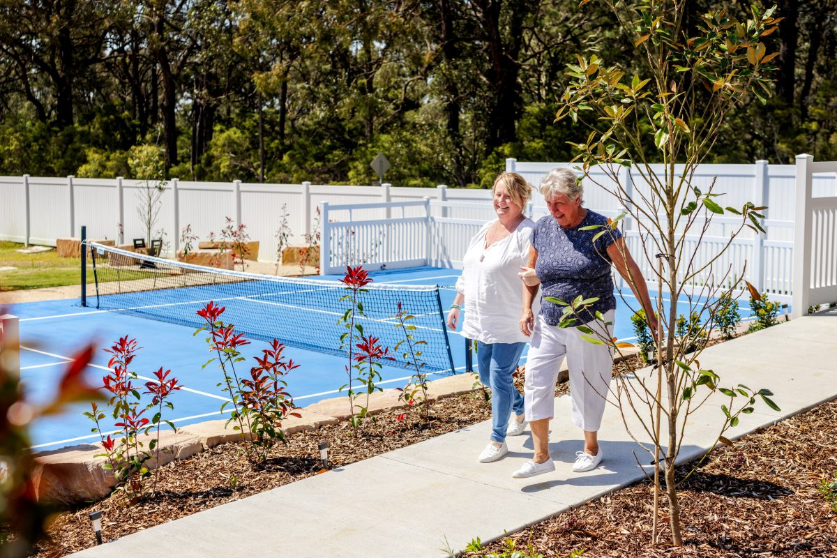 Two older women walk and smile together on a path beside a blue outdoor tennis court, enjoying the relaxed Hunter Valley lifestyle, surrounded by lush plants and trees and a white fence on a sunny day.