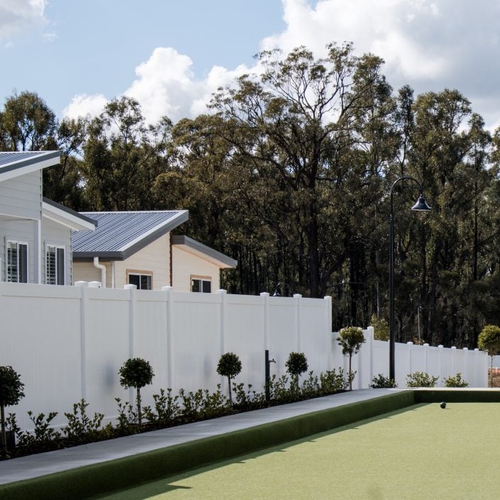 A neatly maintained outdoor lawn bowls green is bordered by a white fence, with modern houses and small shrubs on one side. Enjoy Yoga on the Green in the heart of Hunter, framed by trees under a partly cloudy sky.
