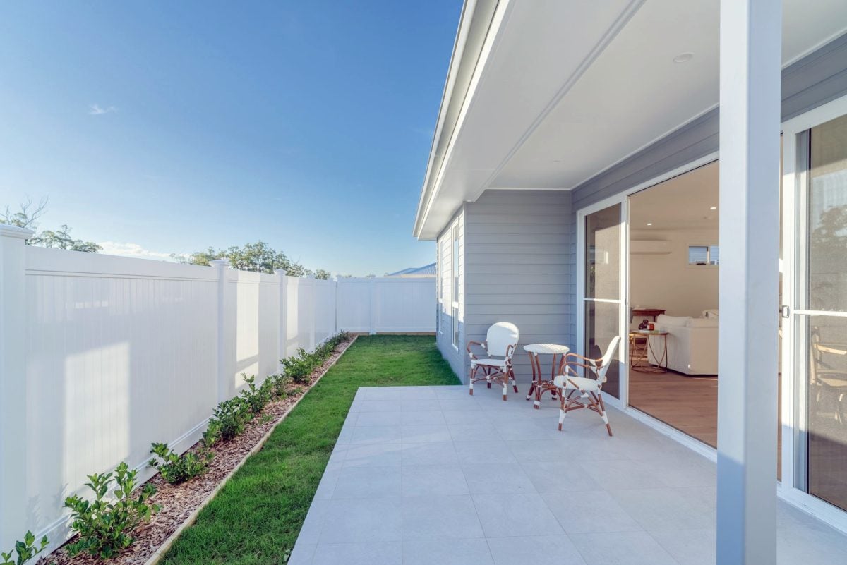 A modern back garden patio with white fencing, light grey cladding, small flower bed, green lawn, and white outdoor furniture showcases thoughtful design next to sliding glass doors leading to a bright living area.