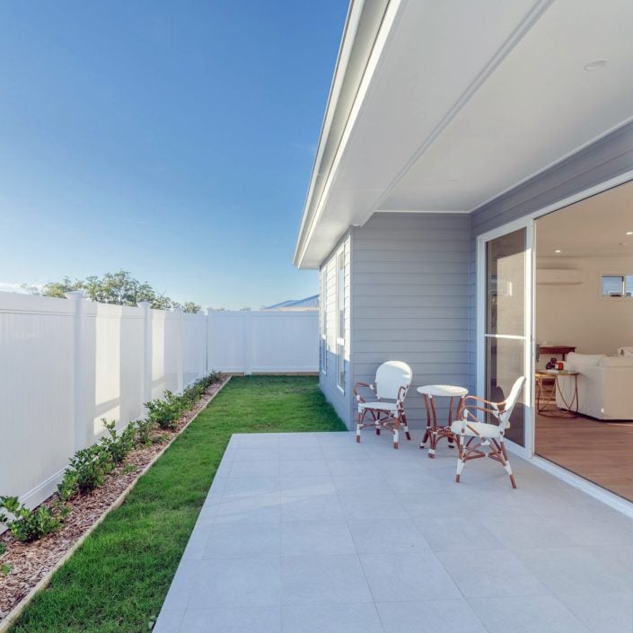 A modern back garden patio with white fencing, light grey cladding, small flower bed, green lawn, and white outdoor furniture showcases thoughtful design next to sliding glass doors leading to a bright living area.