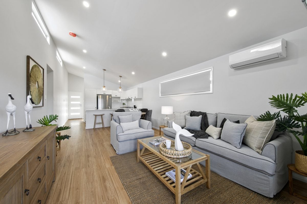 A modern, open-plan living room with light grey sofas, neutral décor, indoor plants evoking Eden Gardens, wooden furniture, and a view into a white kitchen; natural light fills the space.