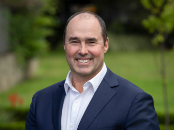 A man in a navy suit and white shirt smiles at the camera whilst standing outdoors in a green, blurred garden setting.