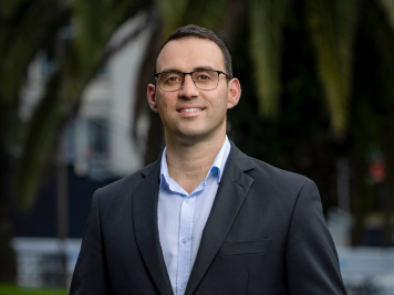 A man wearing glasses, a dark suit jacket, and a light blue shirt is standing outdoors and smiling. There are blurred trees and greenery in the background.