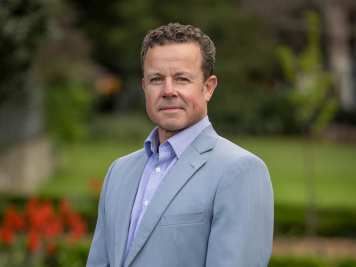 A man in a light grey suit and light purple shirt stands outdoors with a blurred green background and red flowers, looking at the camera with a neutral expression.