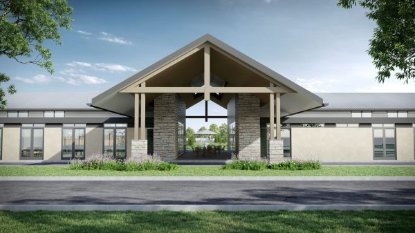A modern club house with a large pitched roof, stone pillars, and wide windows stands behind a road and landscaped greenery in an over-50s lifestyle estate in Mudgee, with trees framing the view under a blue sky.
