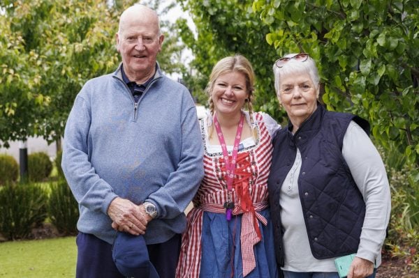 Three adults stand outdoors on a path at Officer Lifestyle Estate. An older man in a blue jumper, a woman in a black waistcoat and sunglasses, and another smiling woman in a traditional blue and red dress are framed by green trees and grass.