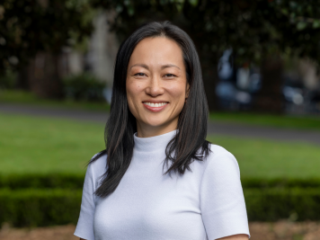 A woman with straight black hair, wearing a white short-sleeve top, smiles whilst standing outdoors in a park with green grass and blurred trees in the background.