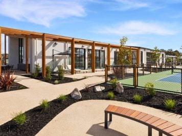 A modern building with large windows and wooden pergolas sits beside a fenced tennis court, offering Huntly’s over-50s inviting landscaped paths, young trees, shrubs, and a curved wooden bench under a blue sky.