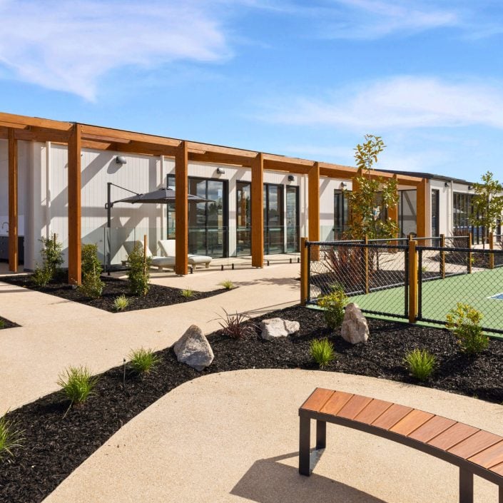 A modern building with large windows and wooden pergolas sits beside a fenced tennis court, offering Huntly’s over-50s inviting landscaped paths, young trees, shrubs, and a curved wooden bench under a blue sky.