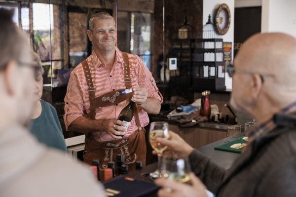 A man in traditional attire opens a wine bottle behind the bar at Officer Lifestyle Estate, smiling at two people in the foreground who hold glasses of white wine at this casual indoor gathering.