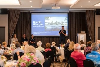 A man speaks on stage at a launch event for Lincoln Lifestyle Northern Beaches, an Over 50s Land Lease Community, with three seated panellists beside him and an audience watching. The presentation slide displays event details for October 2023.