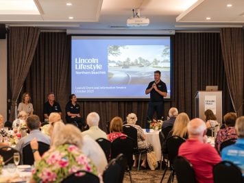 A man speaks on stage at a launch event for Lincoln Lifestyle Northern Beaches, an Over 50s Land Lease Community, with three seated panellists beside him and an audience watching. The presentation slide displays event details for October 2023.