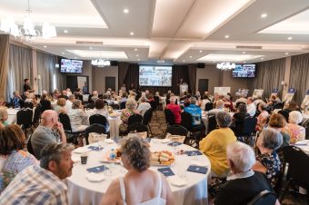A large group of people, mainly Over 50s, sit at round tables in a spacious, well-lit Townsville conference room, watching a presentation. The atmosphere is formal and social, with attendees engaged—typical of a Land Lease Community event.