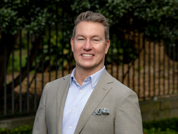 A man with short, fair hair smiles whilst wearing a light grey suit jacket, light blue shirt, and pocket square, standing outdoors in front of greenery and a metal fence.