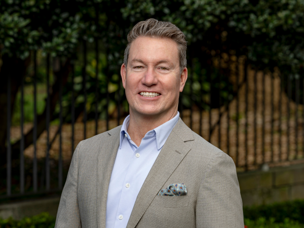 A man with short, fair hair smiles whilst wearing a light grey suit jacket, light blue shirt, and pocket square, standing outdoors in front of greenery and a metal fence.