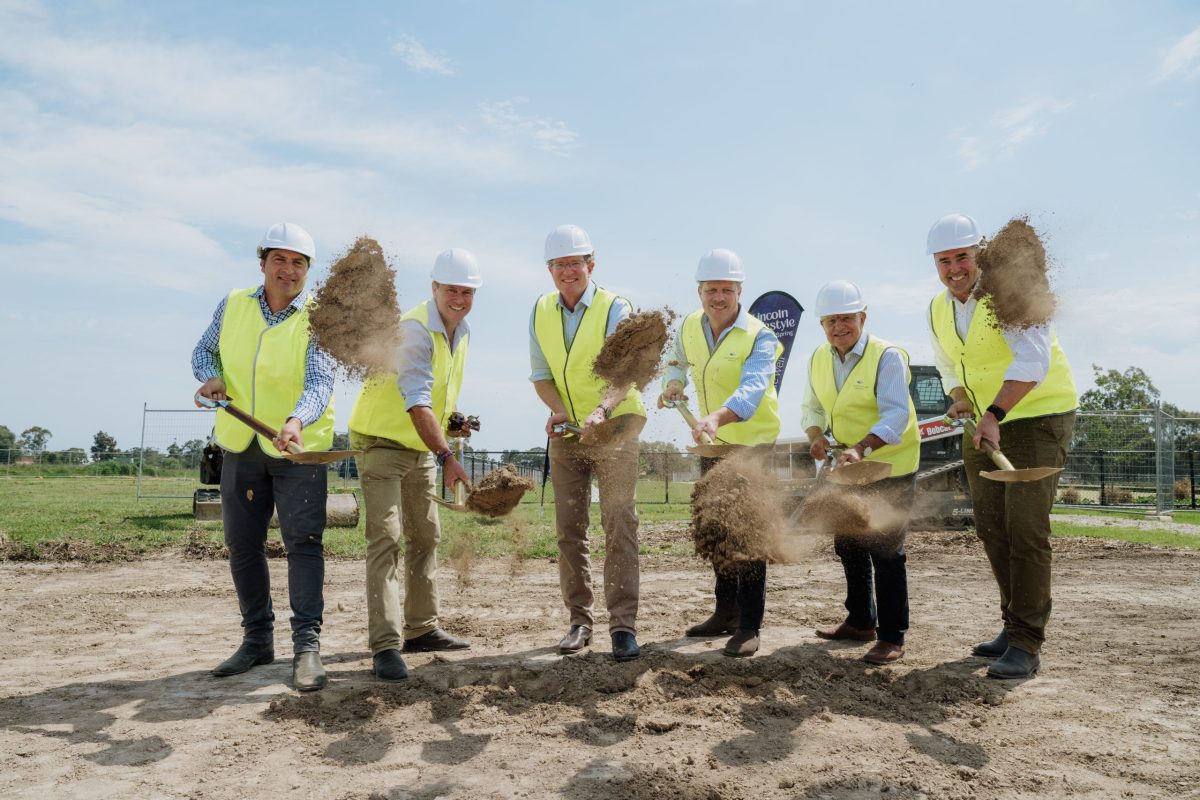 Six people in white hard hats and yellow safety vests smile as they lift shovels of soil at a Mudgee Over 50s lifestyle estate clubhouse groundbreaking ceremony under a clear blue sky.