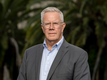 An older man with grey hair and glasses wearing a grey suit jacket and light blue shirt stands outdoors, with green foliage blurred in the background.