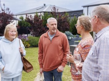 Four older adults stand outdoors in a garden at Officer Lifestyle Estate, chatting and smiling. One woman holds a handbag, another has a glass of wine. The group is casually dressed, with houses and lush plants in the background.