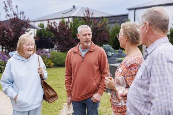 Four older adults stand outdoors in a garden at Officer Lifestyle Estate, chatting and smiling. One woman holds a handbag, another has a glass of wine. The group is casually dressed, with houses and lush plants in the background.