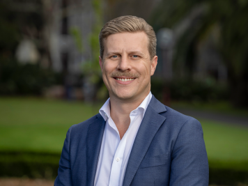 A man with fair hair and a moustache, wearing a blue suit jacket and white shirt, stands outdoors smiling with trees and greenery blurred in the background.