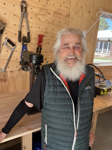 An older man with white hair and a beard smiles whilst standing in The Shed’s wooden workshop. Tools hang on the wall, equipment lines the workbench, and sunlight pours in as residents gather to build bonds over shared projects.