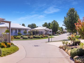 A quiet suburban street with neatly maintained single-storey houses, well-kept lawns, shrubs, and trees under a clear blue sky on a sunny Stage 9 Launch day.