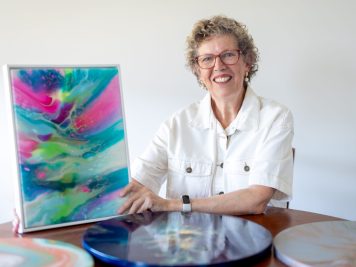 Sylvia, an older woman with curly grey hair and glasses, smiles whilst holding a colourful abstract painting. She sits at a table displaying vibrant, round art pieces, reflecting the creative freedom found at Campbell Lifestyle Estate.