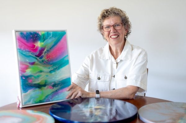 Sylvia, an older woman with curly grey hair and glasses, smiles whilst holding a colourful abstract painting. She sits at a table displaying vibrant, round art pieces, reflecting the creative freedom found at Campbell Lifestyle Estate.