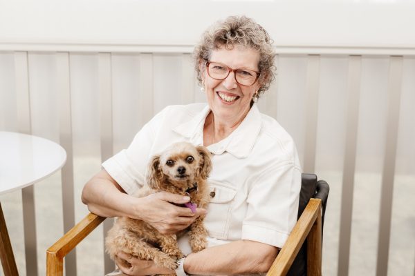 Sylvia, at Campbell Lifestyle Estate, sits in a wooden chair with a smile, embracing her passions as she holds a small, fluffy tan dog on her lap.