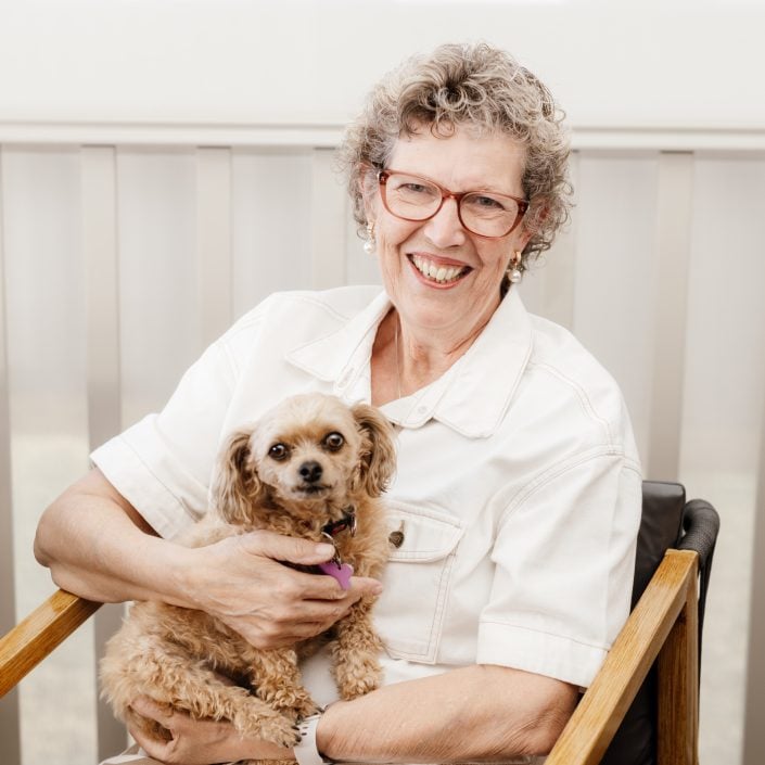 Sylvia, at Campbell Lifestyle Estate, sits in a wooden chair with a smile, embracing her passions as she holds a small, fluffy tan dog on her lap.