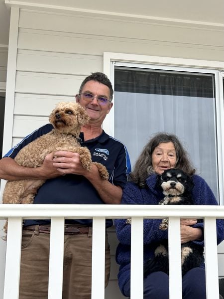 Residents of Eden Gardens, a man and woman stand on a porch holding two small dogs. The man wears glasses and a dark shirt, while the woman wears a blue jacket. Behind them is a white house with a window and white railing.