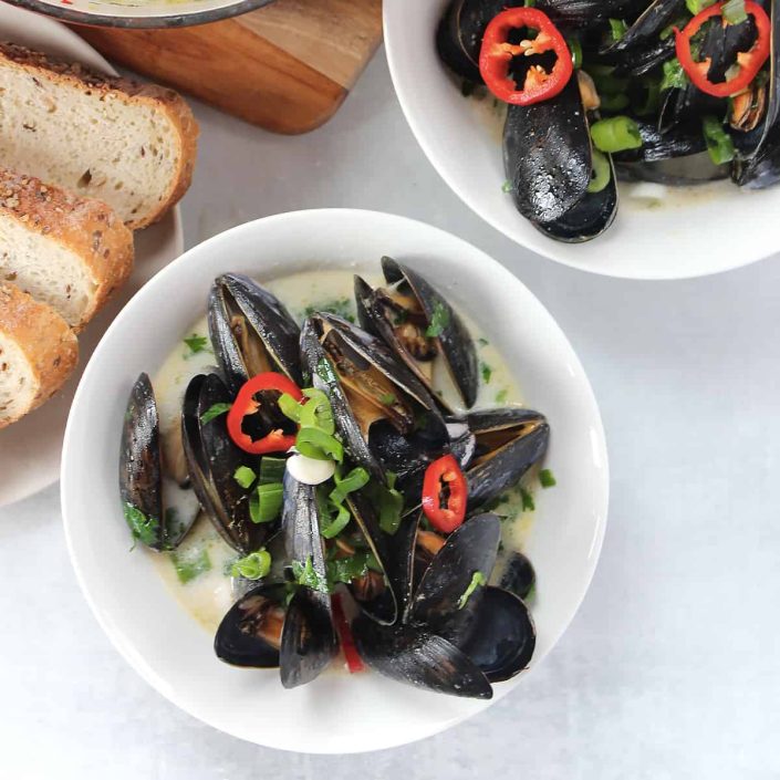 A bowl of steamed mussels in a creamy broth, garnished with sliced red chilli and spring onions, sits next to a plate of bread—an elegant dish perfect for sharing over Lincoln Lifestyle tips.